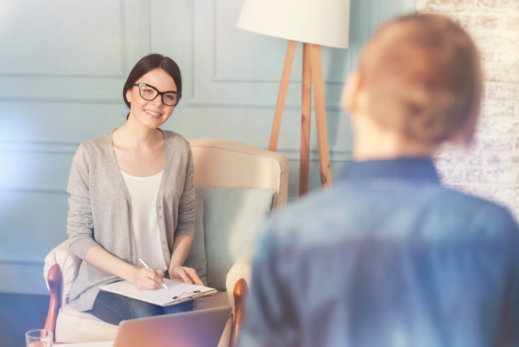 Nice to see you. Cheerful smiling female psychologist sitting on the couch and welcoming a teen boy while going to start a psychological session